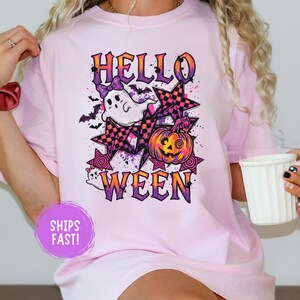 Könnte beinhalten: Ein hellrosa T-Shirt mit einem Grafikdruck und dem Text HELLO WEEN. Das Design zeigt ein weißes Gespenst mit einer lila Schleife, einen orangefarbenen Kürbis, lila-schwarz karierte Sterne und kleine schwarze Fledermäuse auf lila Spritzern.