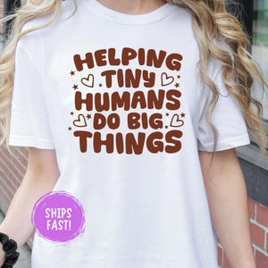 May include: A white short-sleeved t-shirt featuring brown text that reads HELPING TINY HUMANS DO BIG THINGS, accented with small brown heart and star icons. The design is centred on the chest area of the garment, providing a clear graphic display.