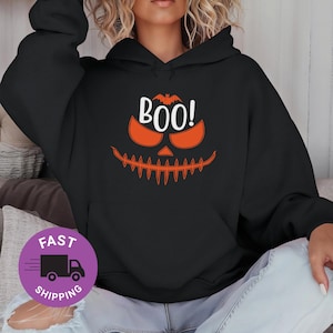 Puede incluir: Sudadera con capucha negra con un diseño temático de Halloween. El diseño presenta una cara de calabaza naranja con la palabra "BOO!" en blanco y un murciélago encima. Un estilo casual y cómodo.