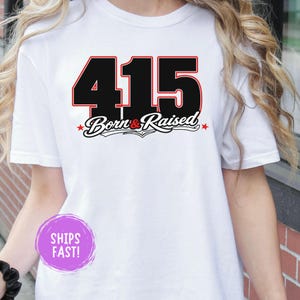 Op de afbeelding: Wit T-shirt met het nummer "415" in zwart met een rode omlijning, en de woorden "Born & Raised" in een scriptlettertype. Het ontwerp bevat rode sterren. Een casual kledingstuk.