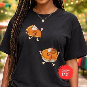May include: A black t-shirt featuring two cartoon pumpkin pie illustrations. The top graphic shows a single slice with whipped cream, while the bottom graphic shows a whole pie with a missing slice, whipped cream, and a small pumpkin garnish.