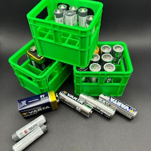 May include: Three green plastic crates stacked on top of each other, filled with various sizes of batteries. The crates have a grid pattern and are designed to hold multiple batteries. The batteries are silver and black and have the brand name "VARTA" printed on them.