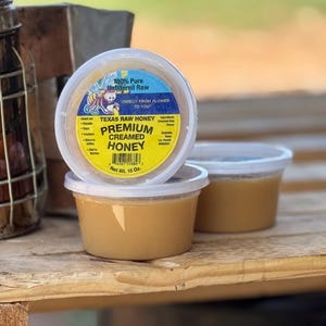 May include: A clear plastic container of Texas Raw Honey Premium Creamed Honey. The container is labeled with the text "100% Pure Unfiltered Raw Direct From Flower To You" and "Texas Raw Honey Premium Creamed Honey". The container is sitting on a wooden surface.