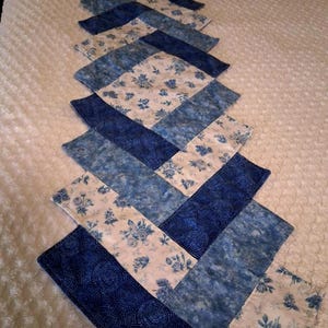May include: A blue and white quilted table runner with a herringbone pattern. The runner features alternating rectangular blocks of solid blue, floral, and patterned fabrics. The floral fabric has a white background with blue roses.