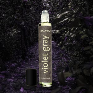 Puede incluir: Un frasco roll-on de vidrio transparente con tapa negra, etiquetado como "Violet Gray". El frasco contiene un l&iacute;quido amarillo claro y se encuentra sobre un fondo oscuro y borroso de follaje. El frasco contiene 10 ml.