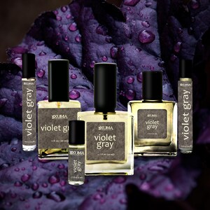 Puede incluir: Varios frascos de perfume con tapas negras y etiquetas blancas con la inscripci&oacute;n "Violet Gray". Los frascos var&iacute;an en tama&ntilde;o, el m&aacute;s grande contiene 50 ml. El fondo es una hoja morada oscura con gotas de agua.