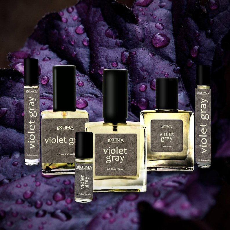 Puede incluir: Varios frascos de perfume con tapas negras y etiquetas blancas con la inscripci&oacute;n "Violet Gray". Los frascos var&iacute;an en tama&ntilde;o, el m&aacute;s grande contiene 50 ml. El fondo es una hoja morada oscura con gotas de agua.