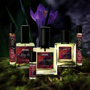 May include: An assortment of perfume bottles with black caps and labels that read "ruby red" in white text. The bottles vary in size, with the largest containing 1.7 fl oz (50 ml). A purple crocus flower is in the background.