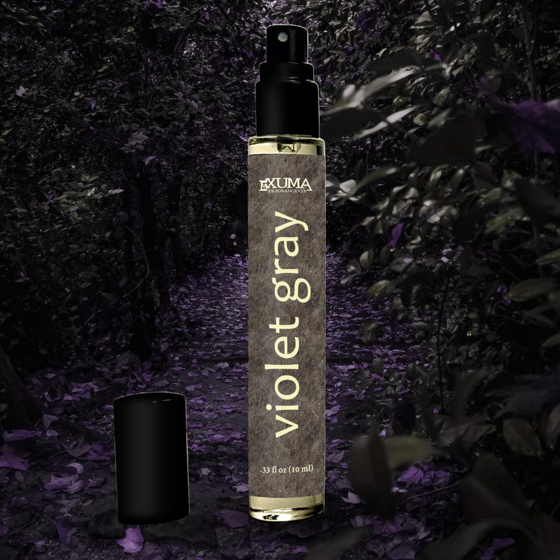 Puede incluir: Un frasco de perfume con tap&oacute;n negro y pulverizador. El frasco est&aacute; etiquetado "Violet Gray" con la marca "EXUMA" y contiene 10 ml. El fondo es una escena borrosa de un bosque con tonos morados.