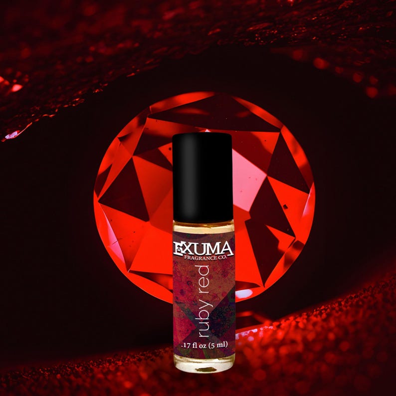 May include: A roll-on perfume bottle with a black cap and a label that reads "ruby red." The bottle is .17 fl oz (5 ml). The background features a large, faceted red gemstone and a dark red, textured surface.