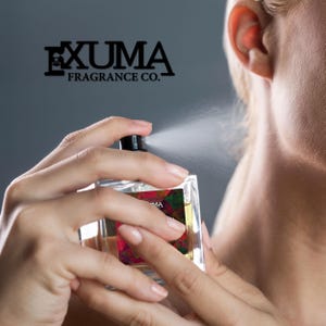 May include: A clear, rectangular perfume bottle being sprayed. The bottle has a black cap and a colorful label. The brand name "EXUMA FRAGRANCE CO." is displayed above the bottle. The perfume is being sprayed towards the neck.