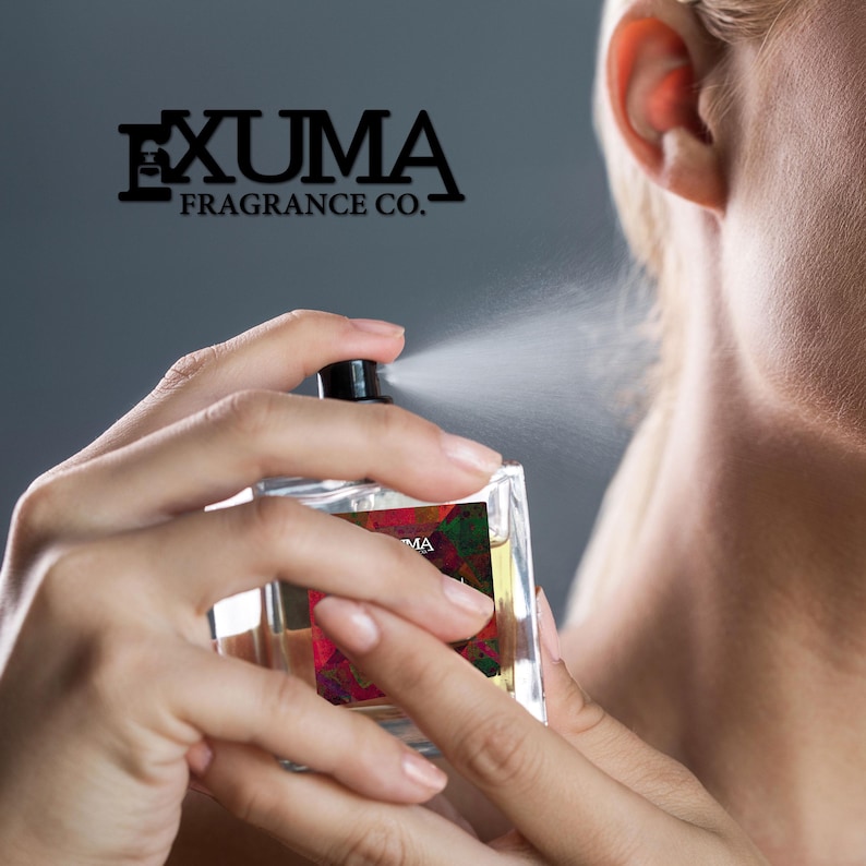 May include: A clear, rectangular perfume bottle being sprayed. The bottle has a black cap and a colorful label. The brand name "EXUMA FRAGRANCE CO." is displayed above the bottle. The perfume is being sprayed towards the neck.