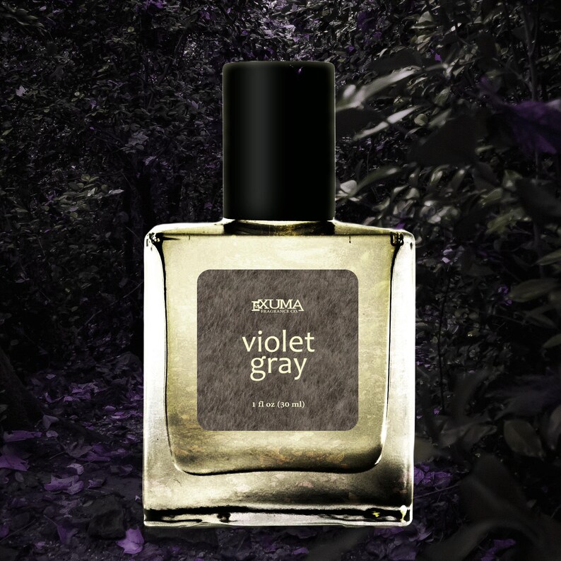 Puede incluir: Botella de perfume cuadrada y transparente con tap&oacute;n negro. La etiqueta gris dice "Violet Gray" y "1 fl oz (30 ml)". El fondo es una imagen oscura y borrosa de follaje. Contiene 30 ml.