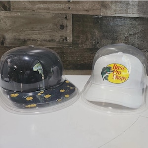 May include: Two baseball caps in clear plastic display cases. One cap is black with a floral pattern on the brim. The other is white with the Bass Pro Shops logo.