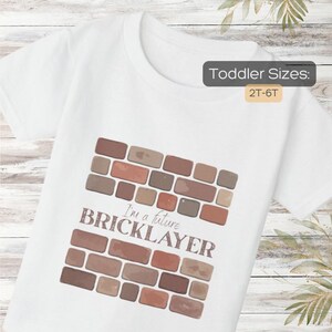 Op de afbeelding: Witte peuter-T-shirt met een bakstenen muurafbeelding. De afbeelding toont een bakstenen muurontwerp in bruin- en tinten, met de tekst "I'm a future BRICKLAYER". Verkrijgbaar in peutermaten 2T-6T.