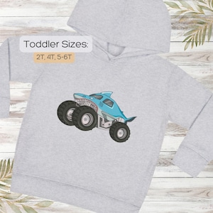 May include: A light gray hooded sweatshirt featuring a blue shark-themed monster truck design. The text "Toddler Sizes: 2T, 4T, 5-6T" is displayed above the graphic. The shark truck has large black tires and a white and red mouth.