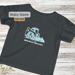 Op de afbeelding: Donkergrijs baby-T-shirt met een lichtblauwe afbeelding van een schranklader en de tekst "I'm a future Skid Steer Operator." Het shirt heeft ook de tekst "Baby Sizes: NB-24M."