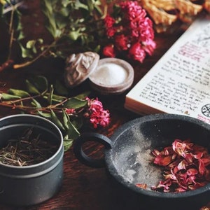 May include: A black cauldron with dried pink flower petals, a glass bottle with dried herbs, a small tin with dried herbs, a book with handwritten text and a pentagram symbol, and a small bowl with white powder on a wooden surface.