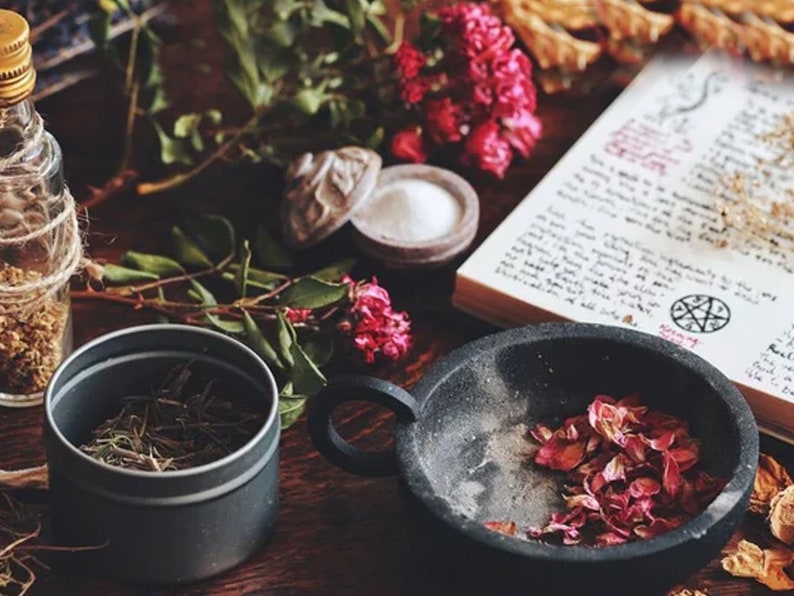 May include: A black cauldron with dried pink flower petals, a glass bottle with dried herbs, a small tin with dried herbs, a book with handwritten text and a pentagram symbol, and a small bowl with white powder on a wooden surface.