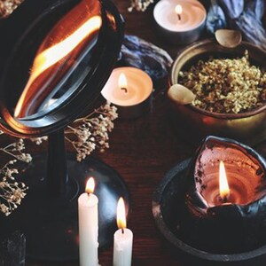 May include: A black vanity mirror with a silver reflective surface, surrounded by candles and dried flowers. The candles are lit and casting a warm glow.