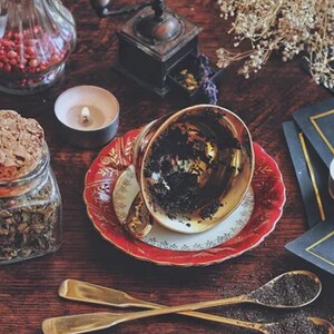 May include: A gold teacup with a red and gold saucer sits on a wooden table. The teacup is empty and has dark tea stains on the inside. There are two candles, a jar of loose leaf tea, and a small glass jar with red berries on the table.