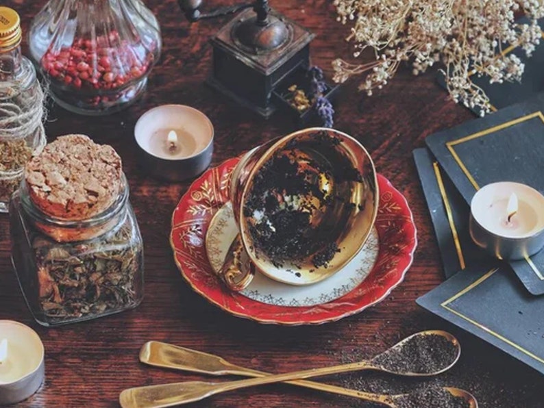 May include: A gold teacup with a red and gold saucer sits on a wooden table. The teacup is empty and has dark tea stains on the inside. There are two candles, a jar of loose leaf tea, and a small glass jar with red berries on the table.