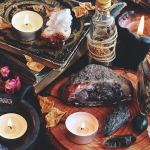 May include: A collection of items for spell casting, including candles, crystals, herbs, and a book. The items are arranged on a wooden surface with a dark background.