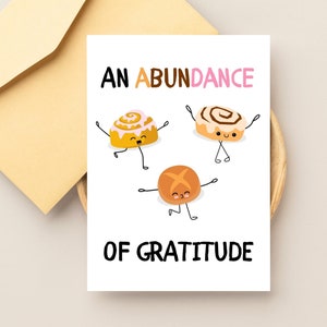 May include: A white card with the text "An Abundance of Gratitude" in black. Three cartoon cinnamon rolls with happy faces are dancing on the card.