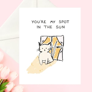 May include: A white greeting card with the text "YOU'RE MY SPOT IN THE SUN". The card features a whimsical illustration of a white cat sitting in front of a window with yellow sunlight and gold stars. A white envelope is visible.