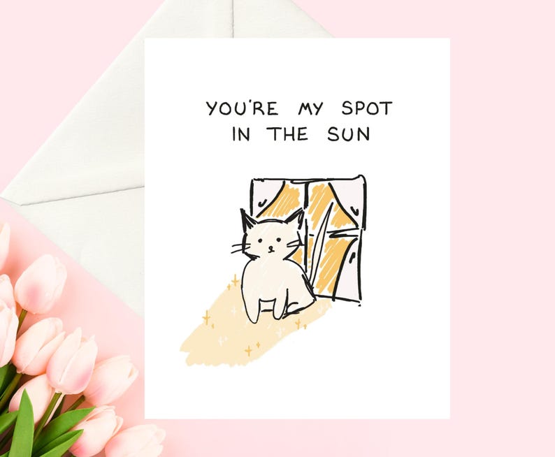 May include: A white greeting card with the text "YOU'RE MY SPOT IN THE SUN". The card features a whimsical illustration of a white cat sitting in front of a window with yellow sunlight and gold stars. A white envelope is visible.