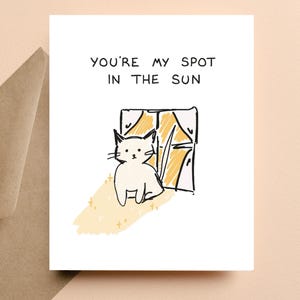 May include: A white greeting card with the text "YOU'RE MY SPOT IN THE SUN" above a sketched illustration of a white cat sitting by a window. The window has yellow curtains and the cat is in a patch of yellow sunlight.