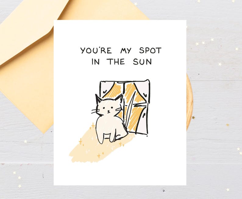May include: A white greeting card with the text "YOU'RE MY SPOT IN THE SUN" and a drawing of a white cat sitting by a window. The card is paired with a light yellow envelope.
