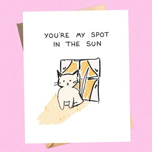 May include: A greeting card with the text "YOU'RE MY SPOT IN THE SUN" above a drawing of a white cat sitting by a window. The window is drawn with yellow and black lines, and the cat is drawn with black lines.
