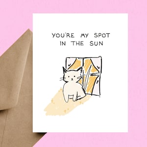 May include: A greeting card with a hand-drawn illustration of a white cat sitting in front of a window with yellow sunlight. The text "YOU'RE MY SPOT IN THE SUN" is at the top. A brown envelope is visible in the background.