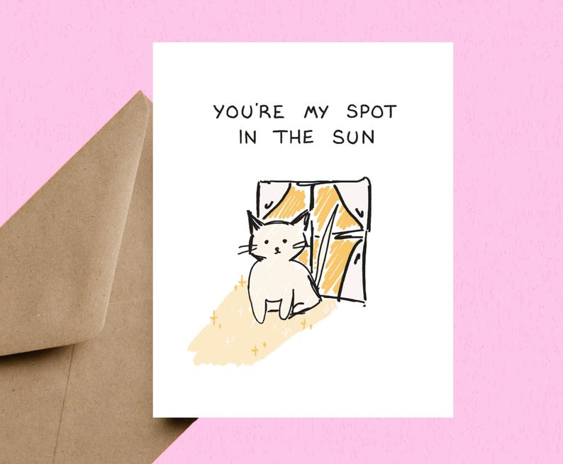 May include: A greeting card with a hand-drawn illustration of a white cat sitting in front of a window with yellow sunlight. The text "YOU'RE MY SPOT IN THE SUN" is at the top. A brown envelope is visible in the background.