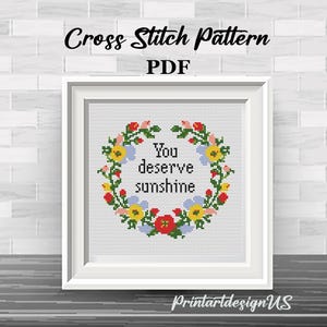 May include: A framed cross-stitch pattern with the text "You deserve sunshine" surrounded by a floral wreath. The frame is white, and the background is a brick wall. The text "Cross Stitch Pattern PDF" is at the top.