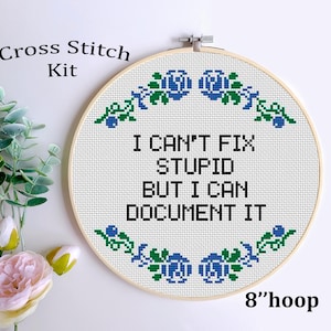 May include: A cross-stitch kit with a 20 cm hoop. The white fabric features the text "I CAN'T FIX STUPID BUT I CAN DOCUMENT IT" in black. The design includes blue and green floral accents. The words "Cross Stitch Kit" are at the top.