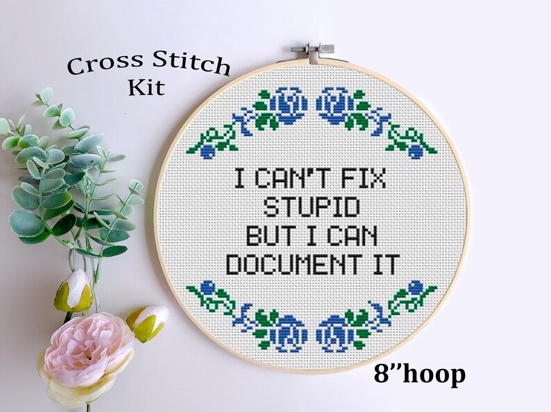 May include: A cross-stitch kit with a 20 cm hoop. The white fabric features the text "I CAN'T FIX STUPID BUT I CAN DOCUMENT IT" in black. The design includes blue and green floral accents. The words "Cross Stitch Kit" are at the top.