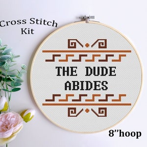 May include: Cross stitch kit featuring the phrase "The Dude Abides" in brown and black lettering on a white background. The design is framed in a brown and orange geometric pattern. The kit is displayed in an 8-inch hoop.