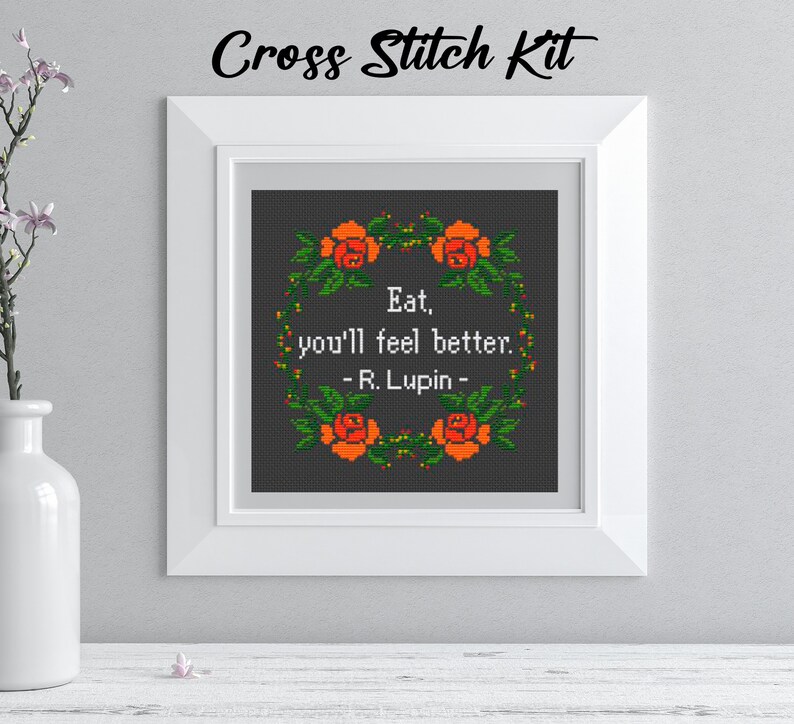 Pu&ograve; includere: Kit per punto croce con un design incorniciato. Il design presenta la citazione "Eat, you'll feel better. - R. Lupin -" all'interno di una ghirlanda di rose arancioni e foglie verdi su sfondo grigio scuro. Le parole "Cross Stitch Kit" sono in alto.