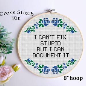 May include: A cross-stitch kit with the text "I CAN'T FIX STUPID BUT I CAN DOCUMENT IT" in black on white fabric. The design is framed in a 20 cm hoop and decorated with blue and green floral accents. The words "Cross Stitch Kit" are at the top.