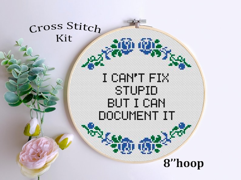 May include: A cross-stitch kit with the text "I CAN'T FIX STUPID BUT I CAN DOCUMENT IT" in black on white fabric. The design is framed in a 20 cm hoop and decorated with blue and green floral accents. The words "Cross Stitch Kit" are at the top.