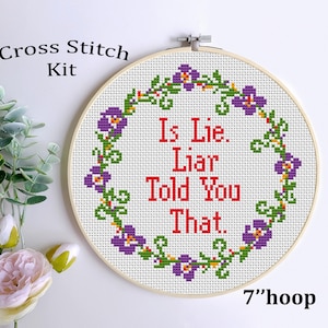 May include: A cross stitch kit in a 7-inch hoop. The design features the text "Is Lie. Liar Told You That." in red, surrounded by a floral border with purple flowers and green leaves. The words "Cross Stitch Kit" are above the hoop.