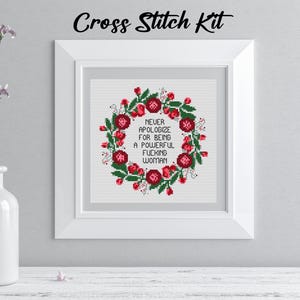 May include: A framed cross-stitch kit with the text "Never apologize for being a powerful fucking woman." The design features a floral wreath in red and green. The words "Cross Stitch Kit" are written above the frame.