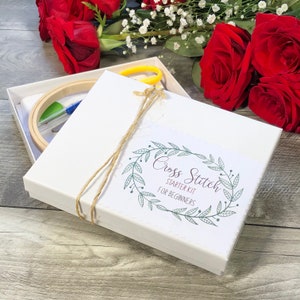 May include: A white box tied with twine, containing a cross stitch starter kit. The kit includes a wooden embroidery hoop, needles, scissors, and fabric. The box has a label that reads "Cross Stitch Starter Kit for Beginners."