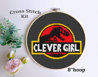 May include: Cross stitch kit featuring a black dinosaur silhouette inside a red circle with a yellow outline. The text "CLEVER GIRL" is below the dinosaur in white letters. The design is on a black fabric in an 8-inch hoop.
