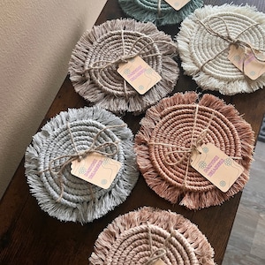 May include: Six round woven coasters with fringe edges in various colors, including beige, light brown, light pink, light blue, and green. Each coaster has a small tag attached with the text "Daisydoe Treasures".