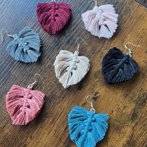May include: Six pairs of handmade macrame earrings in various colors, including red, pink, blue, gray, black, and beige. Each earring features a heart-shaped design with fringe details.