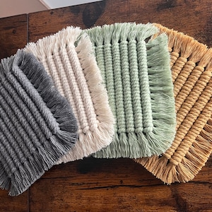 May include: Four square macrame coasters in grey, cream, green, and tan. Each coaster has a woven centre with a fringed edge. The coasters are arranged on a dark wooden surface.