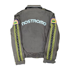 May include: Olive green jacket with the word "NOSTROMO" in light blue across the back. The sleeves feature a vertical design with a dark blue base and light green lacing. Patches with a rainbow and landscape are on the shoulders.
